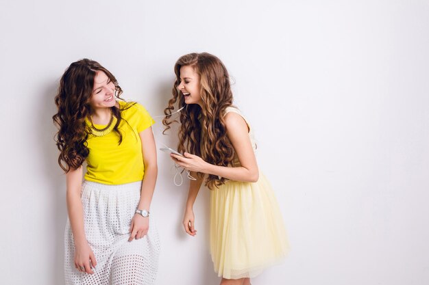 Photo de studio de deux filles souriantes debout écoutant de la musique sur un smartphone et s'amusant. Les filles rient. La brune porte une jupe blanche et un t-shirt jaune, et la fille blonde porte une robe jaune.