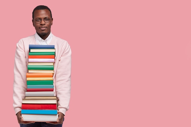 Photo gratuite photo de studio d'un étudiant masculin sérieux à la peau sombre porte une énorme pile de livres, lit et bourre pour l'examen d'entrée