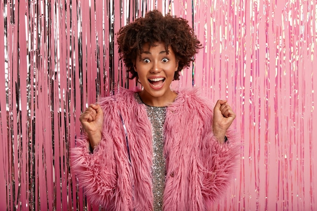 Photo gratuite photo de studio d'une femme afro-américaine optimiste serre les poings, a une réaction heureuse en entendant sa chanson préférée à la fête