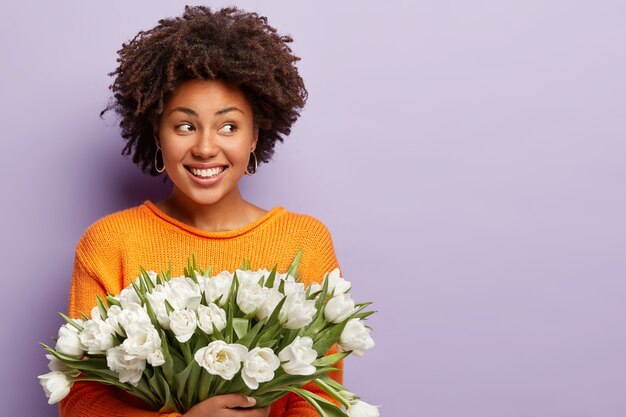 Photo de Studio de femme afro heureuse avec des cheveux croquants, sourit joyeusement, regarde de côté, vêtue d'un pull orange