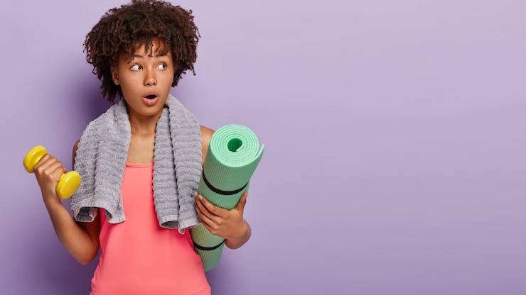 Photo gratuit photo de studio d'une femme afro surprise fait des exercices avec haltères, entraîne l'endurance, utilise un tapis de fitness, vêtue de vêtements de sport