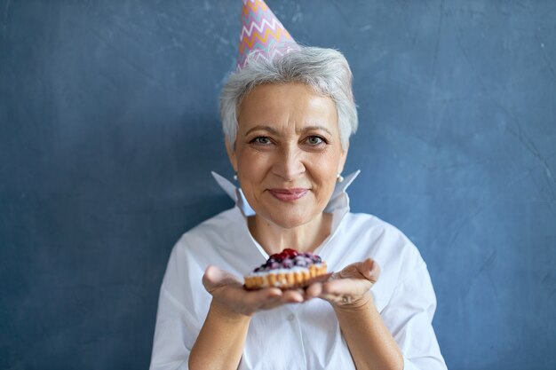 Photo de Studio de femme d'âge moyen beauitful heureux portant un chapeau conique célébrant l'anniversaire, posant isolé avec un gâteau dans ses mains, vous offrant d'avoir mordu. Mise au point sélective sur le visage de la femme