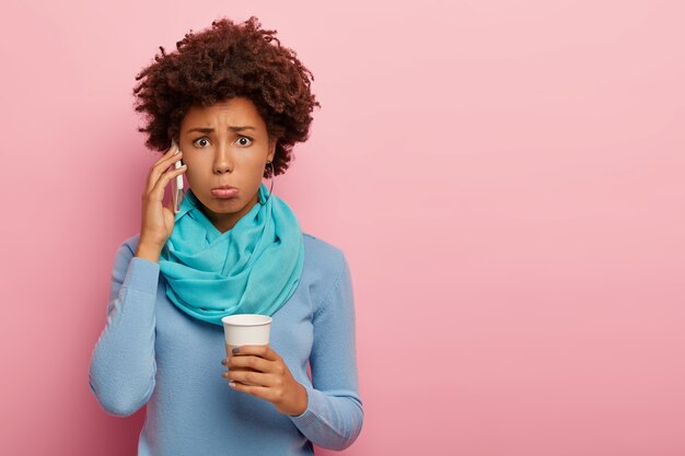 Photo de Studio d'une femme bouclée insatisfaite tient le téléphone mobile près de l'oreille, a surpris le regard malheureux, boit du café, porte des vêtements décontractés bleus, pose sur fond rose