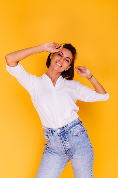 Photo de Studio de femme heureuse cheveux courts portant chemise blanche et pantalon en denim posant sur mur jaune