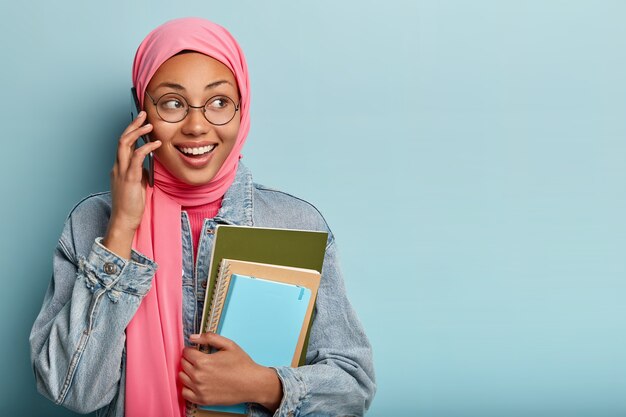Photo de Studio d'une femme positive heureuse a des opinions religieuses islamiques, parle de quelque chose sur un téléphone intelligent