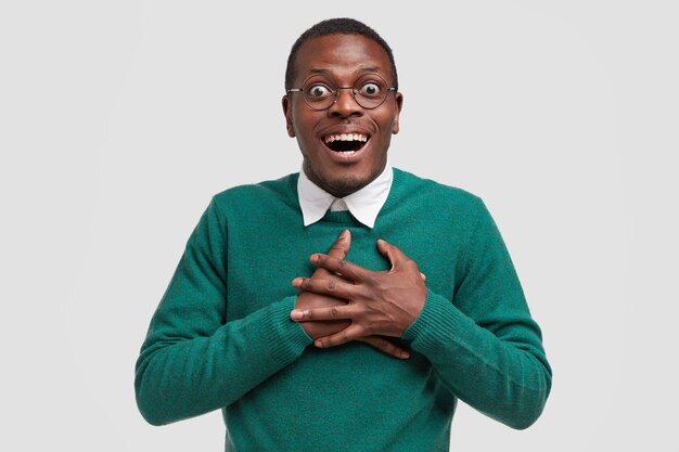 Photo de Studio d'un homme gai regarde avec étonnement et bonheur, garde les mains sur la poitrine, vêtu d'un pull vert