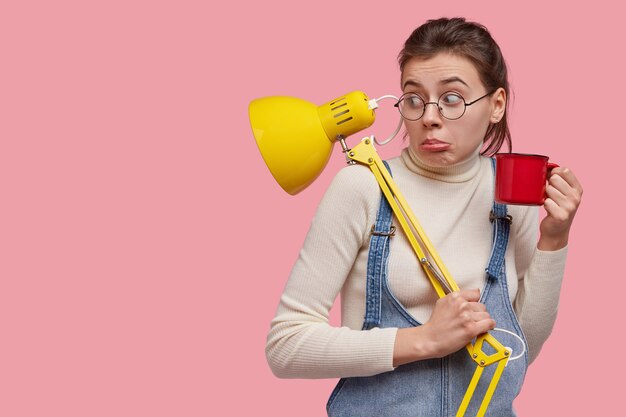 Photo de Studio de jeune adolescent perplexe porte-monnaie lèvre inférieure, regarde étonnamment de côté, porte des lunettes rondes, a les cheveux noirs, remarque quelque chose de désagréable