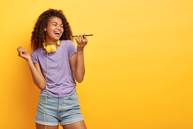 Photo de Studio de joyeuse jeune femme avec une coiffure afro posant contre le mur jaune