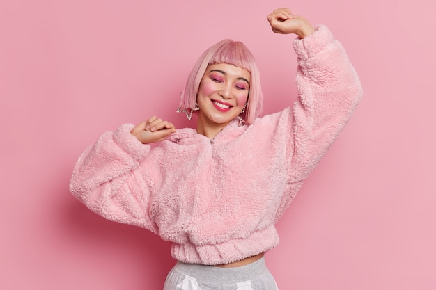 Photo de Studio de joyeuse jeune jolie femme asiatique porte perruque rose maquillage lumineux soulève les bras se sent des danses optimistes sans soucis célèbre quelque chose habillé en manteau de fourrure