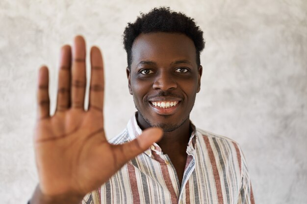 Photo de Studio de joyeux jeune homme africain souriant largement à la caméra, faisant panneau d'arrêt. Beau mec noir en chemise rayée disant bonjour, ami de voeux, ayant l'air heureux