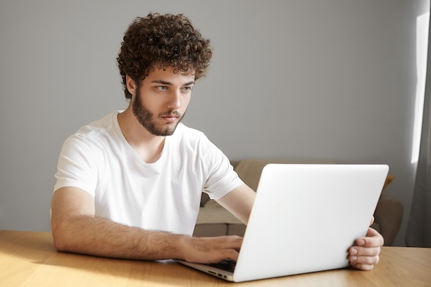 Photo de talentueux jeune rédacteur barbu en t-shirt décontracté tapant sur un ordinateur portable générique dans son intérieur minimaliste de bureau à domicile, regardant l'écran avec une expression concentrée