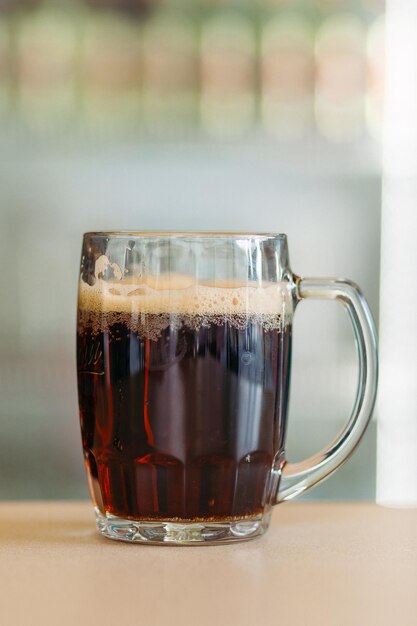 Photo d'une tasse de bière en verre debout sur une surface en bois lisse Boisson d'été froide et sombre pour la chaleur du jour Frais avec une mousse haute et épaisse Arrière-plan flou clair Concept de prise de vue de boissons alcoolisées