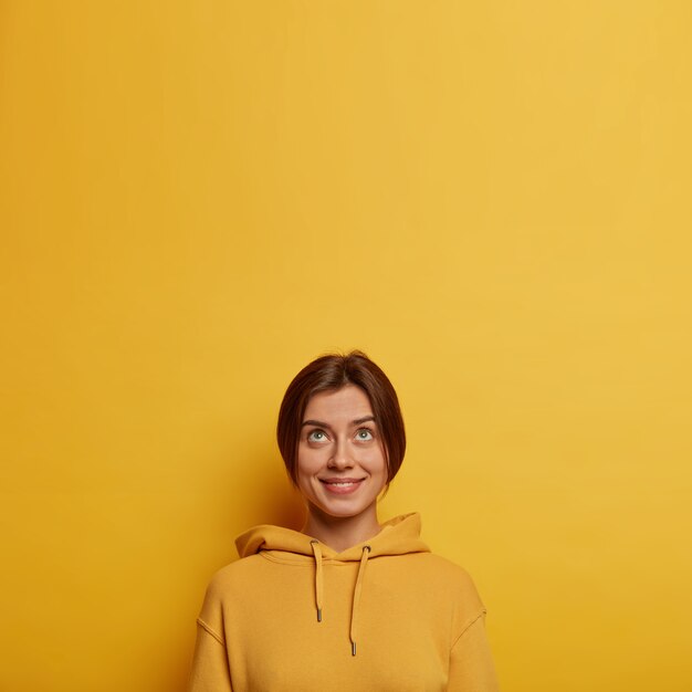 Photo de tendre belle jeune femme vêtue d'un sweat-shirt décontracté, regarde ci-dessus avec intérêt, sourit doucement