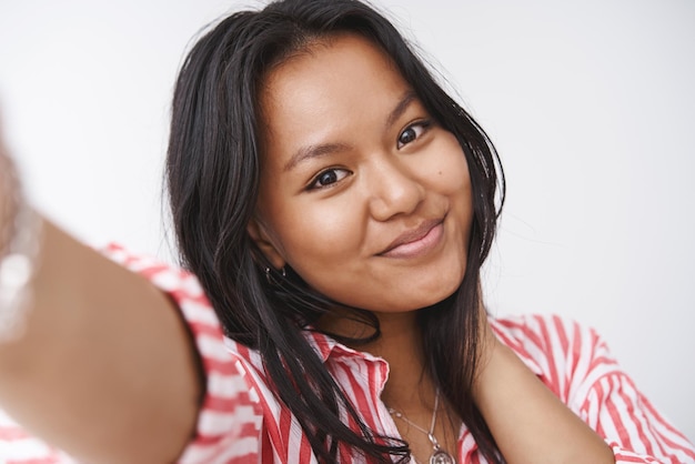 Photo gratuite photo de tête d'une charmante jeune femme polynésienne mignonne et tendre tirant la main vers la caméra en prenant un selfie et en regardant la caméra avec un joli sourire doux posant sur fond blanc