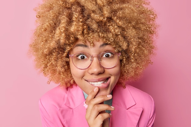 Photo gratuite photo de tête d'une femme curieuse joyeuse mord les lèvres regarde avec joie la caméra écoute de bonnes nouvelles porte des lunettes transparentes et une veste formelle isolée sur fond rose expressions du visage heureux