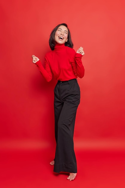 Photo de toute la longueur d'une belle femme brune dansant sans soucis, garde les mains levées, porte un col roulé et un pantalon noir isolé sur fond rouge vif. Modèle féminin énergique et insouciant pose à l'intérieur