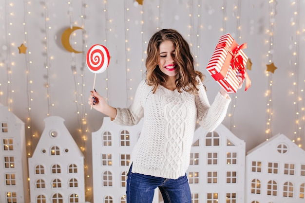 Photo de vacances du nouvel an de jeune femme drôle en pull tricoté doux et jean bleu, dansant avec un cadeau dans sa main gauche et de gros bonbons dans sa main droite
