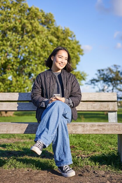 Photo verticale d'une belle femme brune assise sur un banc avec un smartphone attendant quelqu'un utilisant