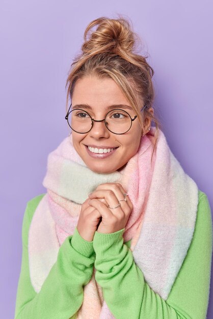 Photo verticale d'une belle femme heureuse sourit joyeusement garde les mains ensemble concentrées à distance pense à quelque chose d'agréable porte des lunettes rondes une écharpe chaude autour du cou rappelle quelque chose