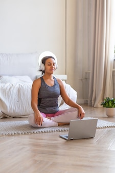 Photo verticale d'une femme afro-américaine médite à la maison devant un écran d'ordinateur portable et écoute de la musique via des écouteurs.