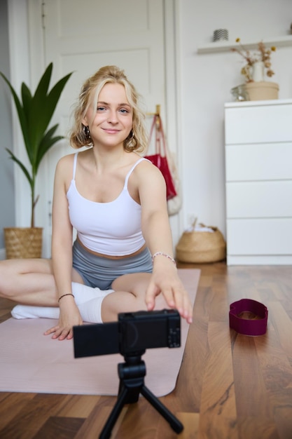 Photo gratuite une photo verticale d'une femme assise sur un tapis de yoga en train d'enregistrer une vidéo sur un appareil photo numérique en faisant un vlog sur l'entraînement.