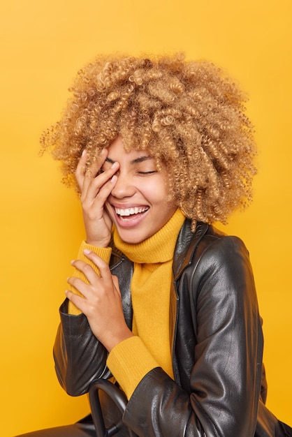 Photo verticale d'une femme aux cheveux bouclés positifs couvre le visage avec les mains sourit largement garde les yeux fermés rit joyeusement porte un pull chaud et une veste en cuir isolés sur fond jaune vif