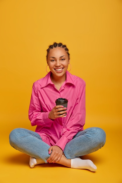 Photo gratuite photo verticale d'une femme heureuse à la peau sombre est assise en posture de lotus, jambes croisées sur le sol, boit du café à emporter, se sent à l'aise, bénéficie d'une conversation amicale avec un interlocuteur, isolé sur un mur jaune