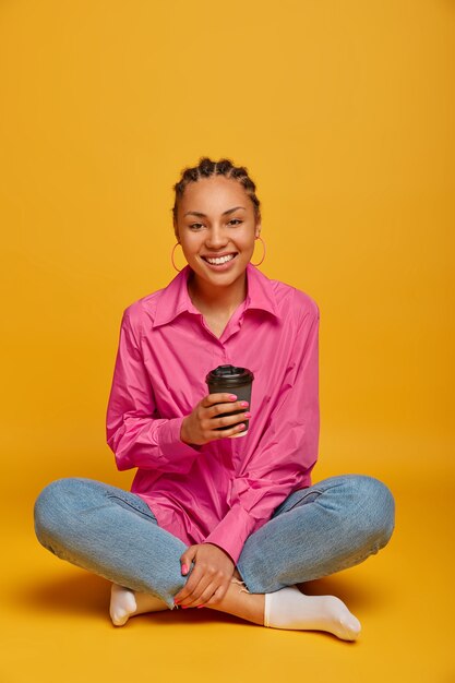 Photo verticale d'une femme heureuse à la peau sombre est assise en posture de lotus, jambes croisées sur le sol, boit du café à emporter, se sent à l'aise, bénéficie d'une conversation amicale avec un interlocuteur, isolé sur un mur jaune