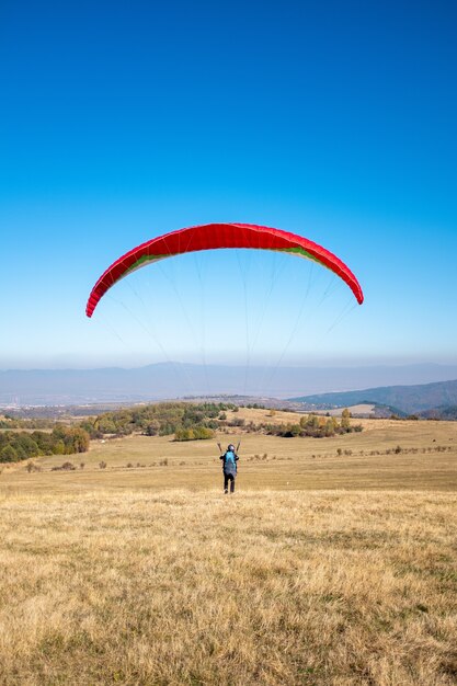 Photo verticale d'un homme volant avec un parachute rouge entouré de verdure sous un ciel bleu