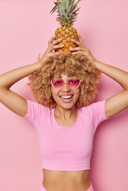 Photo verticale d'une jeune femme enjouée et joyeuse tenant des ananas frais sur la tête porte des lunettes de soleil en forme de coeur recadrées top stupides autour isolé sur fond rose Concept de style de vie d'été