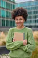 Photo gratuite une photo verticale d'une jeune femme heureuse aux cheveux bouclés tient un bloc-notes et un stylo note ce qu'elle observe dans la ville vêtue d'un pull vert décontracté pose à l'extérieur sur fond flou
