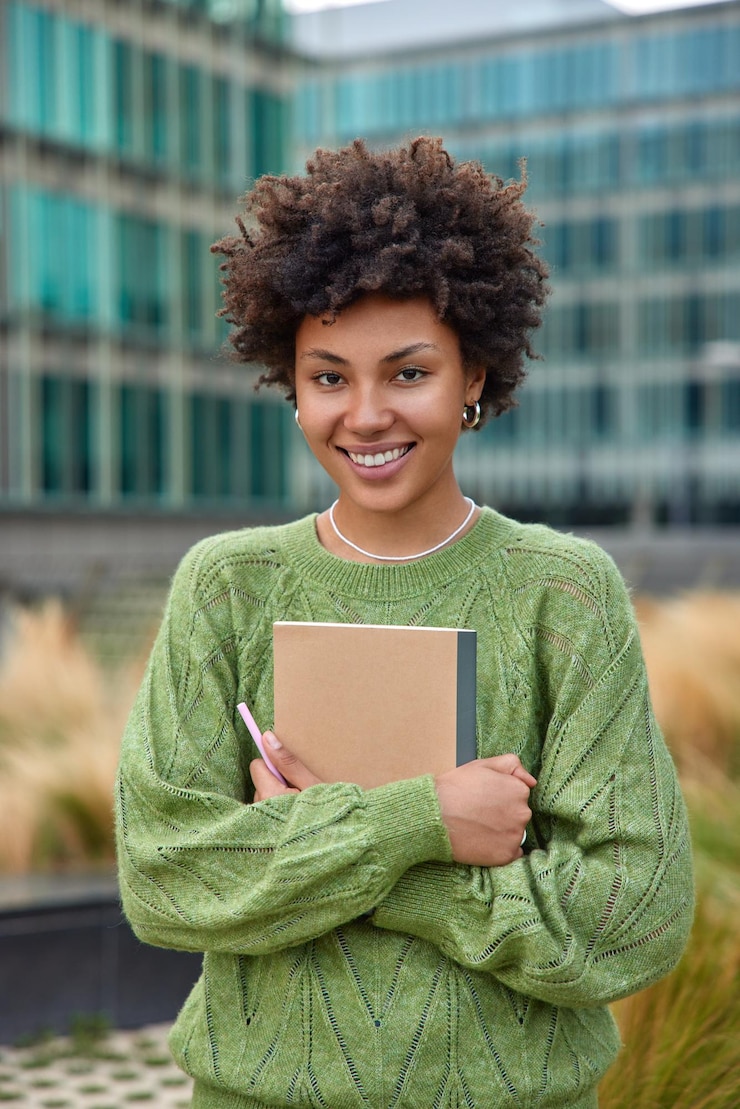 Photo gratuit une photo verticale d'une jeune femme heureuse aux cheveux bouclés tient un bloc-notes et un stylo note ce qu'elle observe dans la ville vêtue d'un pull vert décontracté pose à l'extérieur sur fond flou