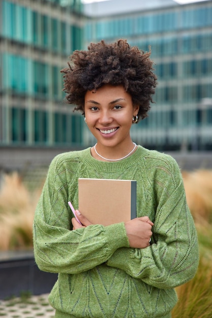 Une photo verticale d'une jeune femme heureuse aux cheveux bouclés tient un bloc-notes et un stylo note ce qu'elle observe dans la ville vêtue d'un pull vert décontracté pose à l'extérieur sur fond flou