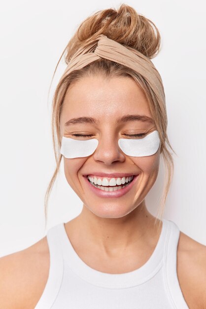 La photo verticale d'une jeune femme heureuse garde les yeux fermés, les sourires applique joyeusement des patchs de beauté sous les yeux, porte un bandeau et un t-shirt isolés sur fond blanc. Concept de soins de la peau sous les yeux.