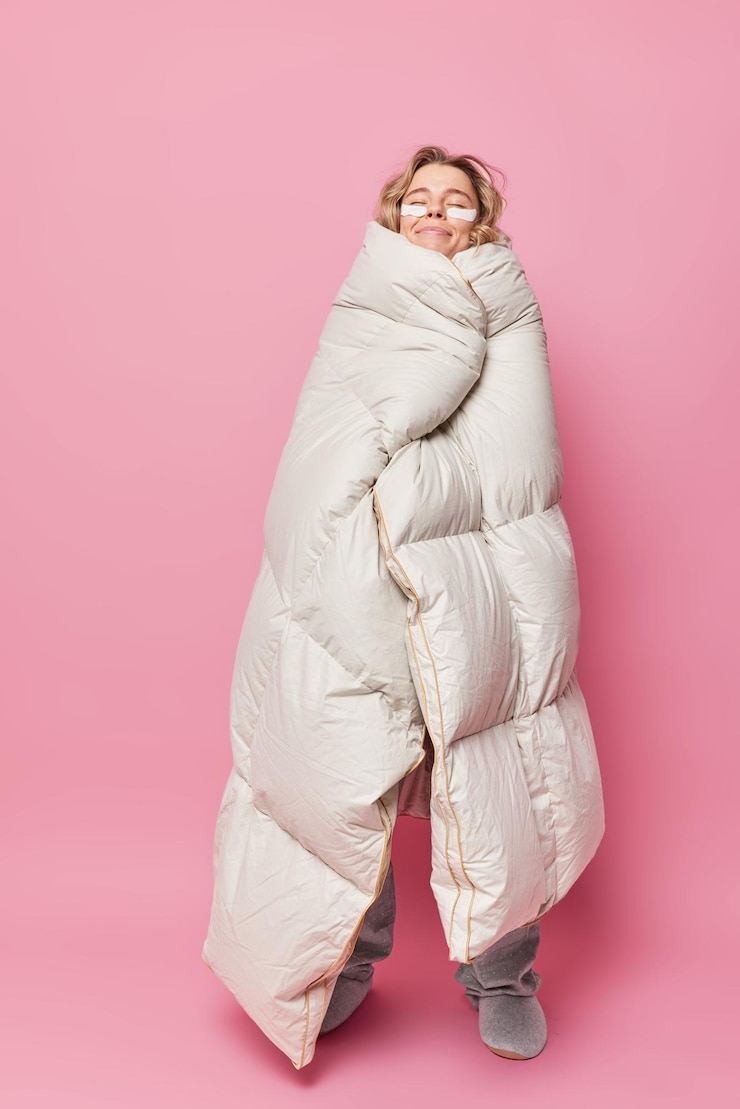 Photo gratuit la photo verticale d'une jeune femme heureuse se sent au chaud alors qu'elle est enveloppée dans des couvertures douces avec les yeux fermés applique des patchs de beauté pose en pleine longueur sur fond rose. heure du matin et réveil