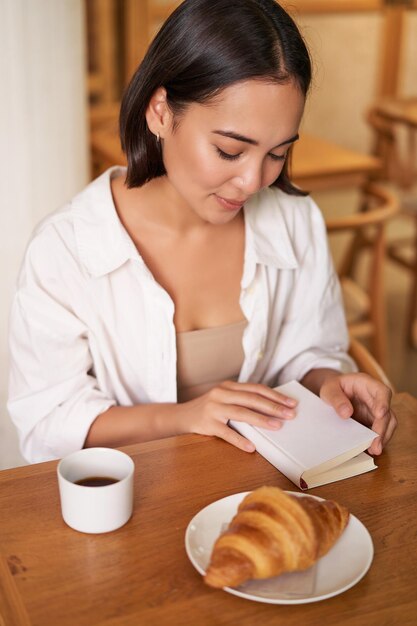 Photo verticale d'une jeune femme souriante avec un livre lisant au café en appréciant le café du petit-déjeuner le matin et