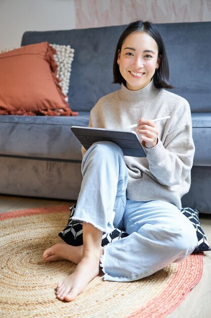 Photo gratuite photo verticale d'une jolie fille asiatique dans une maison confortable s'appuyant sur sa tablette numérique avec un concepteur de stylo workin