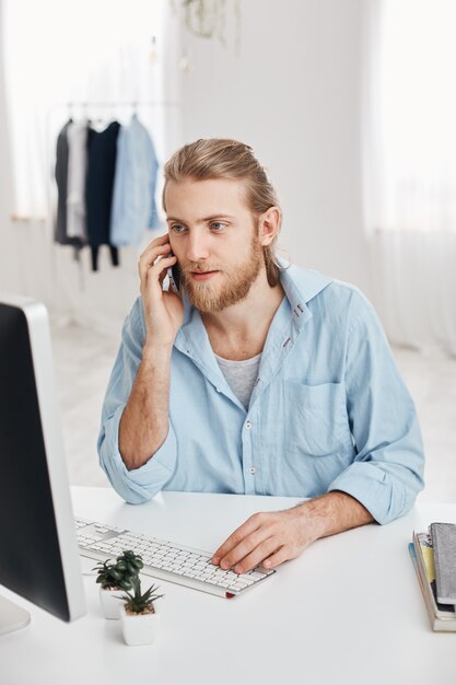 Photo verticale de pigiste beau caucasien avec barbe et cheveux blonds vérifier les informations et taper le texte promotionnel. Un employé masculin agréable a une conversation téléphonique avec des clients.