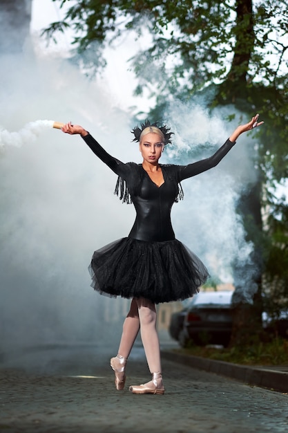 Photo gratuite photo verticale sur toute la longueur d'une belle ballerine aux cheveux blonds portant un corset noir et un tutu dansant dans les rues de la ville fumant sur le fond une performance épique expressive et dramatique.