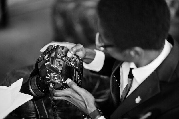 Photographe afro-américain paparazzi homme portant un costume noir et des lunettes assis au bureau avec caméra et travaillant derrière un ordinateur portable