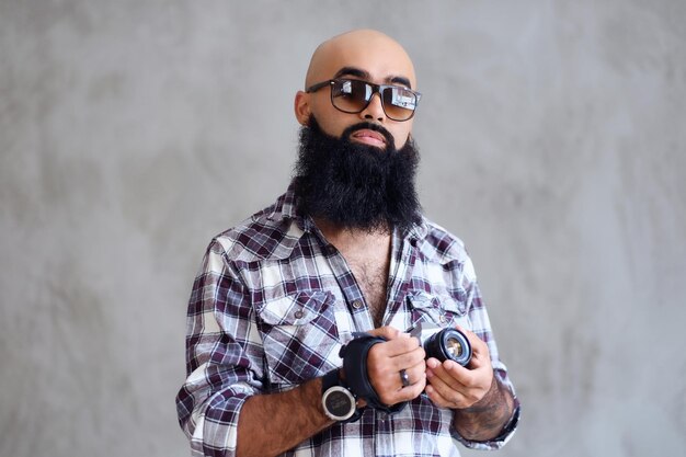 Un photographe amateur hipster barbu avec des tatouages sur les bras, vêtu d'une chemise polaire tient un appareil photo reflex numérique compact sur fond gris.