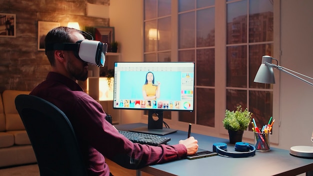Photographe indépendant utilisant un casque de réalité virtuelle tout en retouchant des photos avec une tablette graphique au bureau à domicile pendant la nuit.