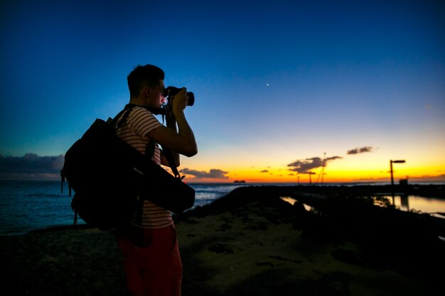 Le photographe se tient debout avec une caméra sur le rivage avec un beau ciel du soir derrière lui