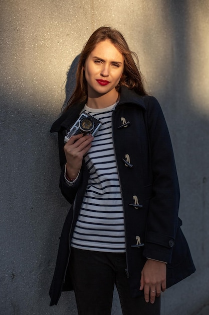 Photographe souriante en veste debout devant le mur prête à faire une nouvelle photo. Adorable jeune femme brune en tenue tendance posant sur fond de mur en béton avec caméra