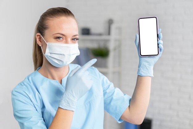 Physiothérapeute femme avec masque médical tenant et pointant sur smartphone