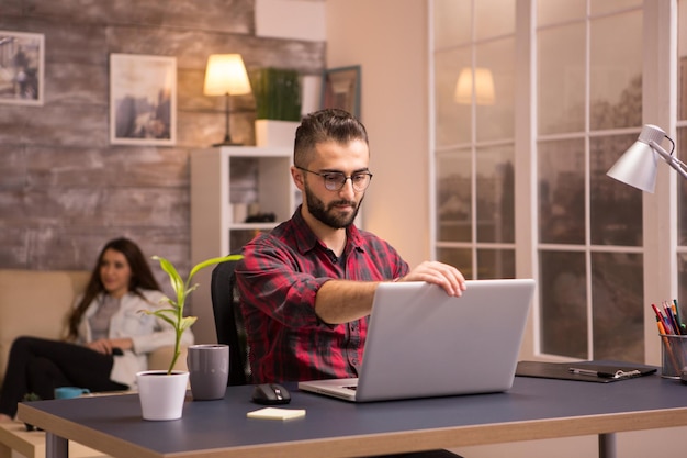 Un pigiste barbu ouvrant son ordinateur portable pour commencer à travailler dans le salon. Petite amie se détendre sur le canapé en regardant la télévision en arrière-plan.