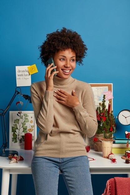 Photo gratuite une pigiste joyeuse aux cheveux bouclés discute avec un collègue, aime la conversation téléphonique, est heureuse d'entendre quelque chose, se sent joyeuse sur un travail à distance, se tient dans son propre cabinet près du lieu de travail.