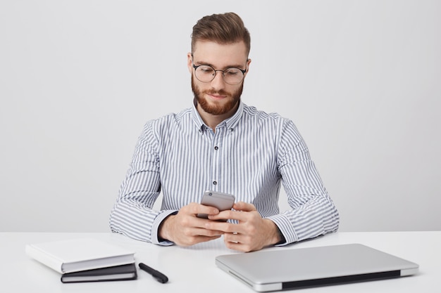 Un pigiste masculin confiant avec une coiffure à la mode travaille à distance, regarde l'écran du téléphone intelligent avec une expression ciblée, communique en ligne, bénéficie d'une connexion Wi-Fi gratuite au bureau, isolé sur un mur blanc