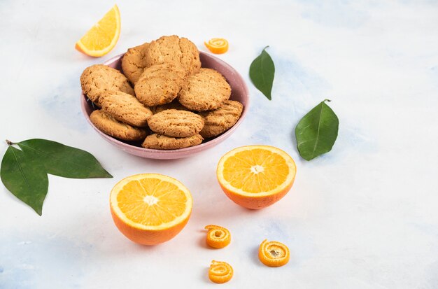 Pile de biscuits dans un bol rose et oranges à moitié coupées avec des feuilles sur une table blanche.