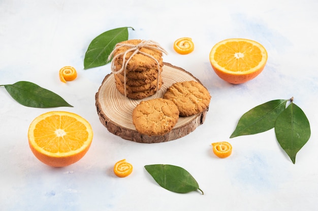 Pile de biscuits faits maison sur planche de bois et orange à moitié coupée avec des feuilles sur une surface blanche.